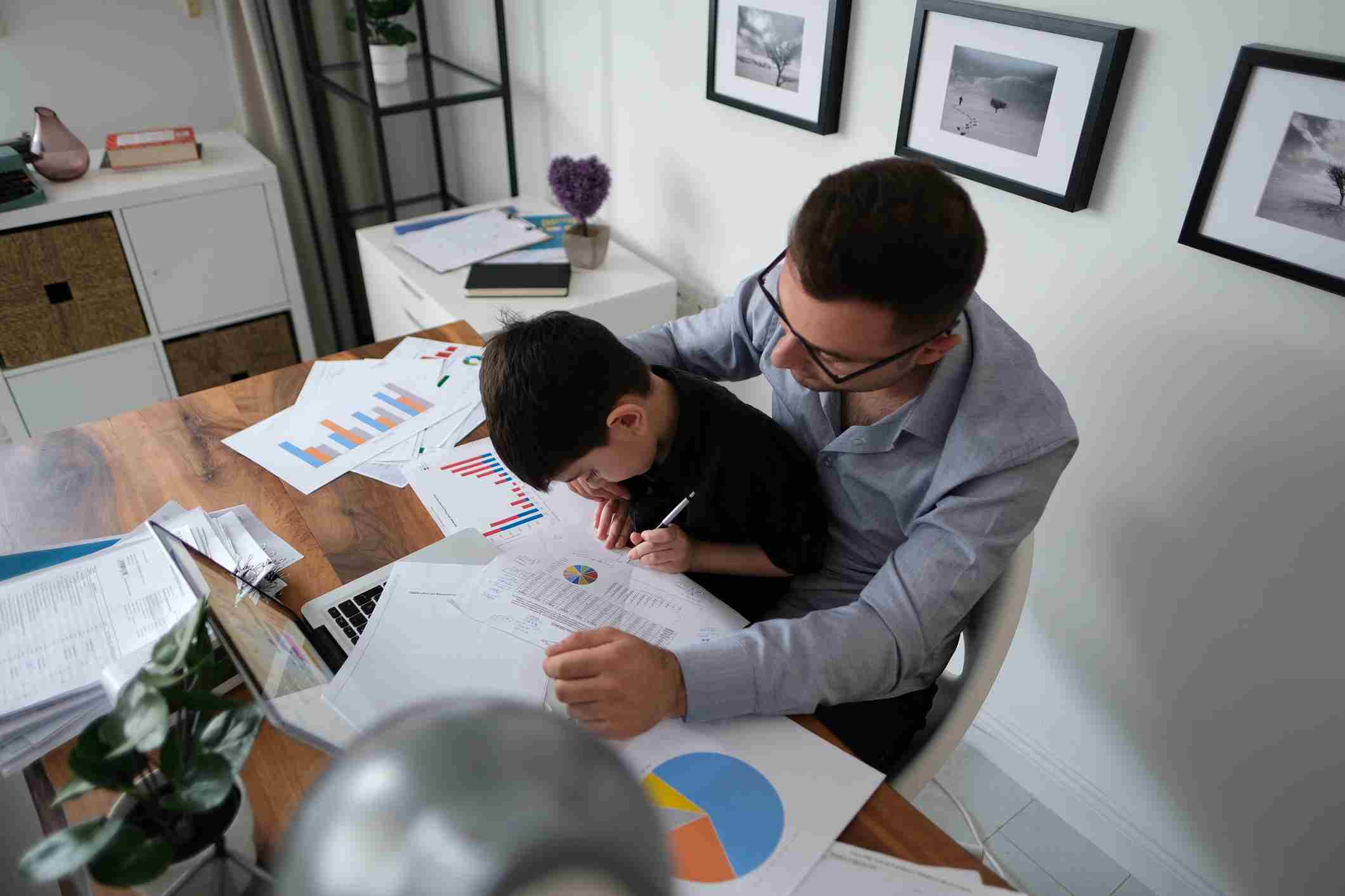 A man helping a boy with homework at a desk covered in papers and charts
