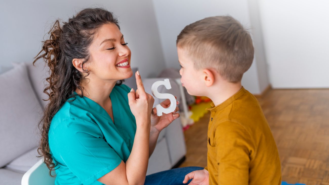 A speech therapist smiling and pointing to her mouth while a young boy watches