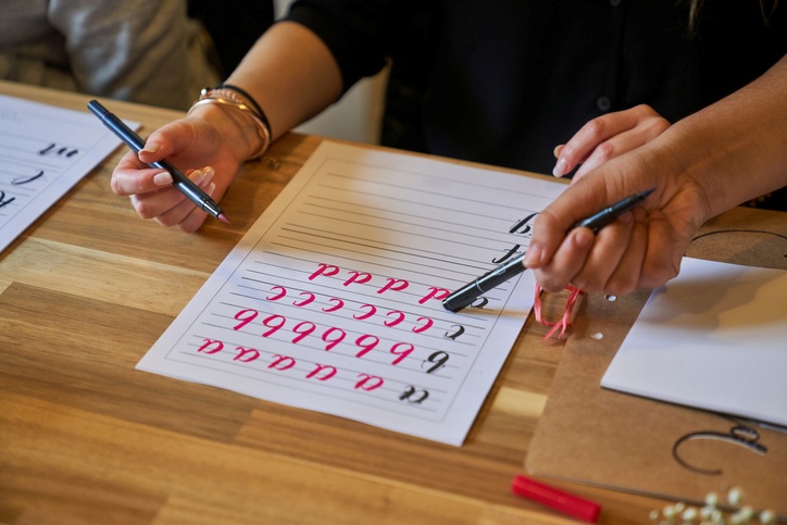 Close-up of two people's hands practicing handwriting letters on a practice sheet
